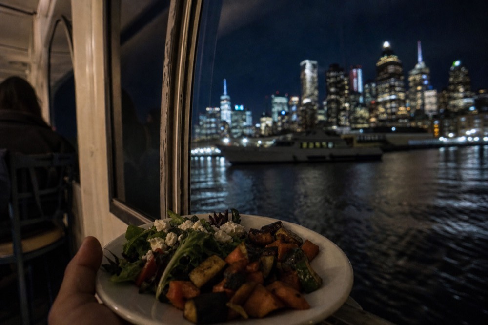 A plate of salads and roasted vegetables held near a boat window with the city skyline out of focus beyond
