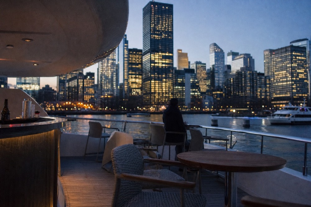 Small upper deck of a yacht with a compact bar and a few seats, city towers rising close behind the rail in evening light