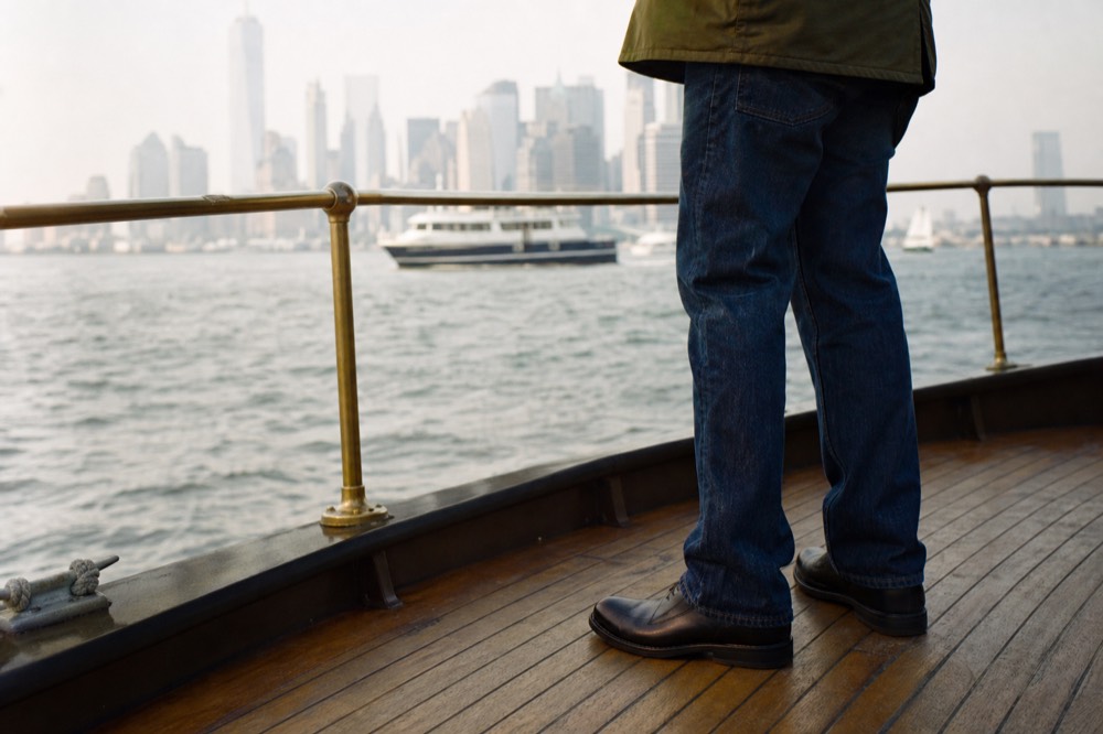 Detail of dark indigo denim with polished leather shoes on a wooden boat deck, brass rail and blurred harbor water behind.