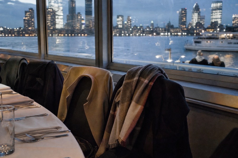 A row of coats and a folded wrap draped over the backs of dining chairs beside a boat window at night, table set with cutlery.