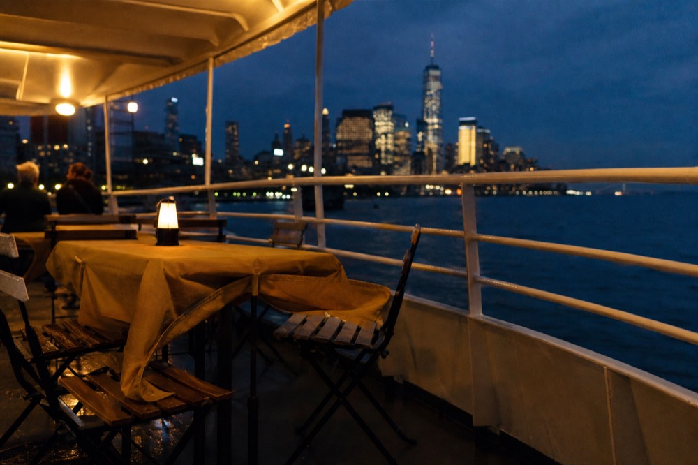 An empty open-air upper deck of a harbor boat in low autumn light, wind rippling a tablecloth, skyline towers in soft focus behind.