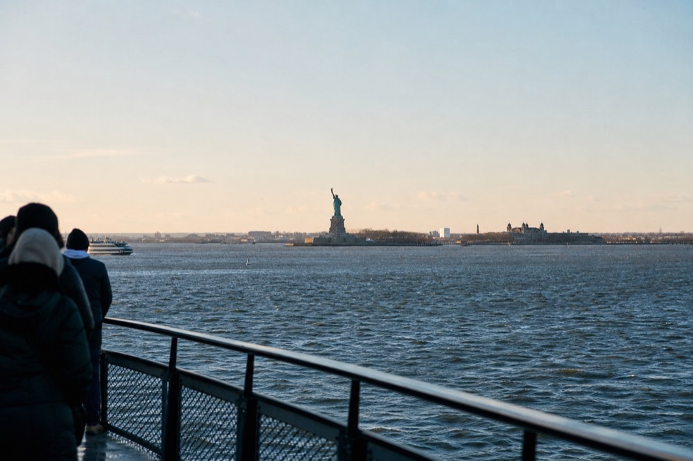 Wide view of Upper New York Bay from a boat deck, the statue small and centred on the water with Ellis Island buildings behind