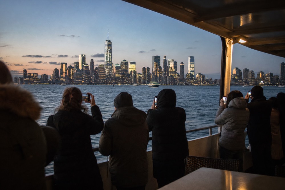Passengers standing at an open boat rail in coats photographing a lit skyline across dark harbor water