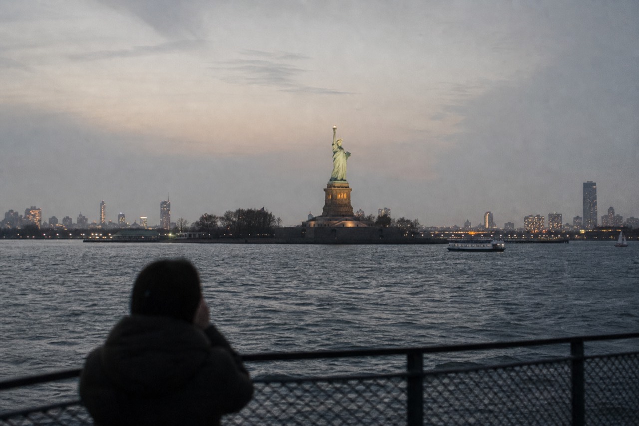 The Statue of Liberty seen across dark water from the rail of a passing harbor boat at dusk, torch lit, city glow behind
