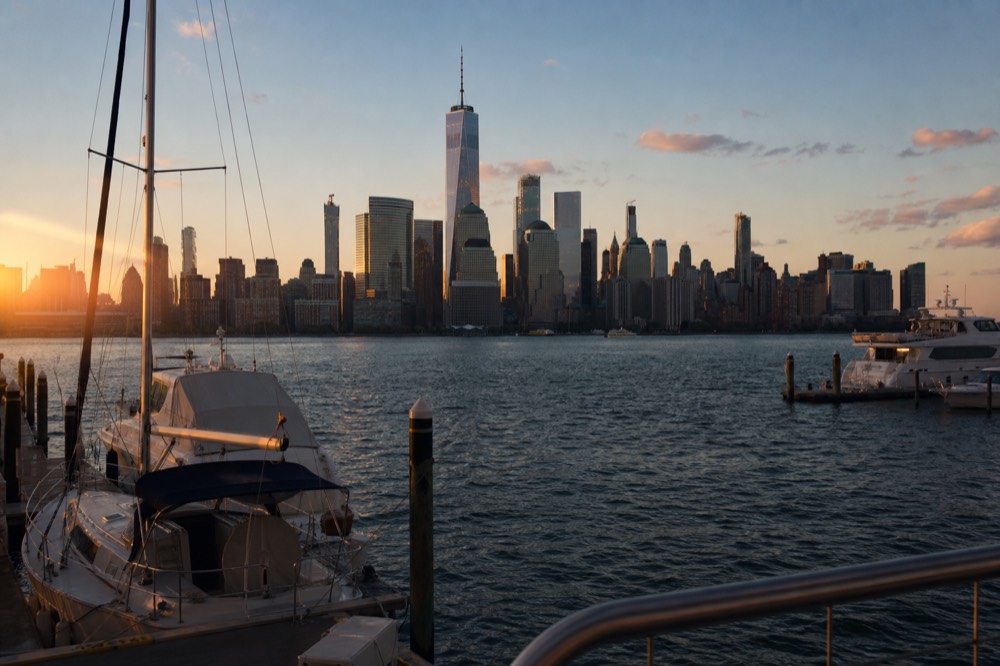 A wide marina basin at dusk with moored boats in the foreground and the full Manhattan skyline standing across the water