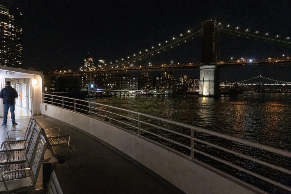 A cruise vessel's open stern deck at night, warm interior light spilling out, the lit Brooklyn Bridge spanning the dark water behind