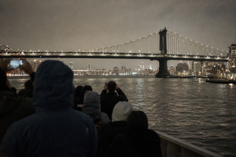 Passengers in coats on a boat deck at night photographing a lit suspension bridge, dark water and orange city glow behind them