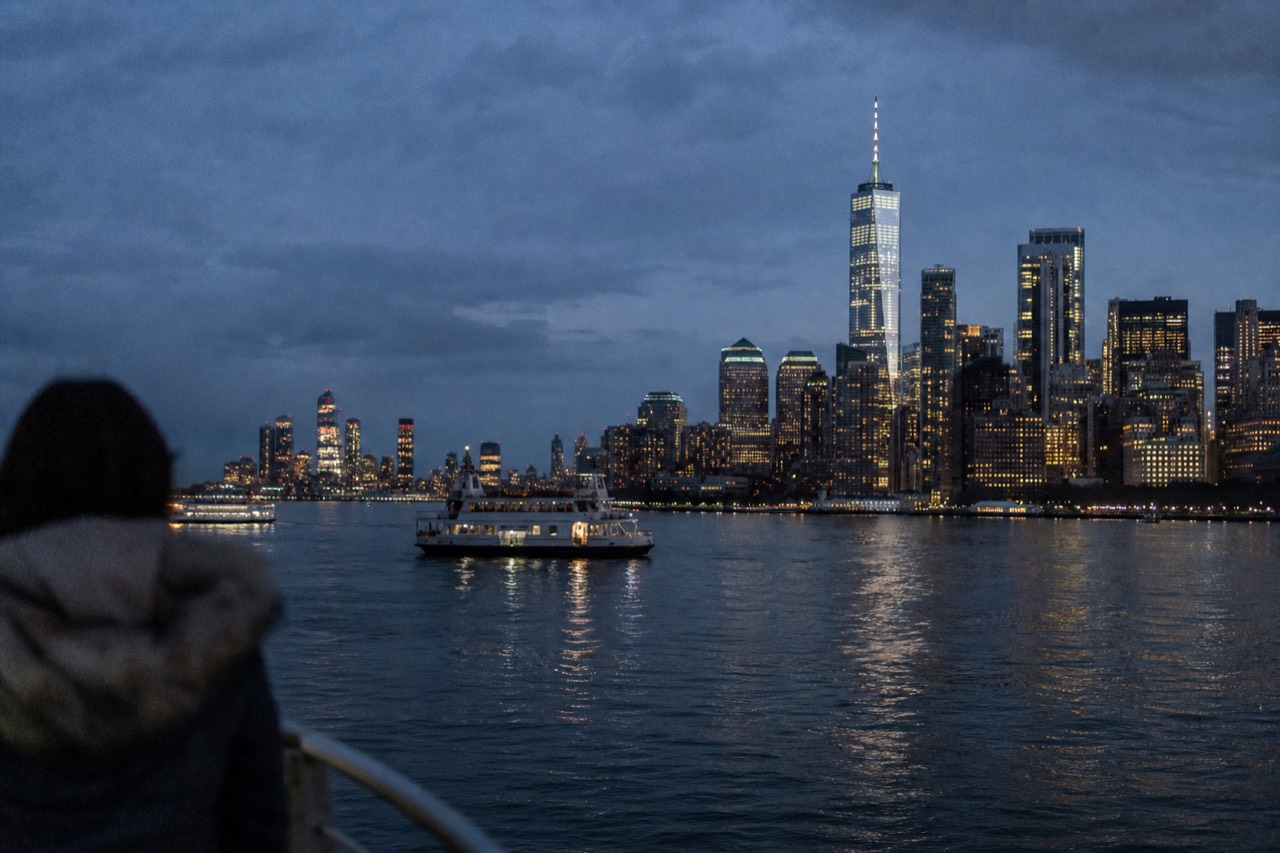 The lit Manhattan skyline seen low from the water at night, tower lights doubling in the black surface of the harbor