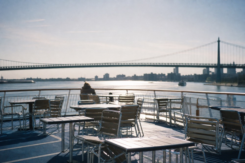 Empty upper deck seating on a cruise boat at midday, low sun on the water, distant bridge span across the frame