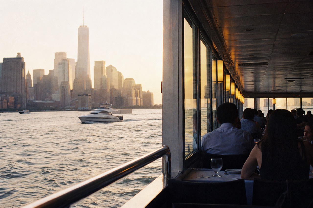 Diners at white-clothed tables beside a wall of glass on a harbor boat, Manhattan towers lit gold behind them at dusk