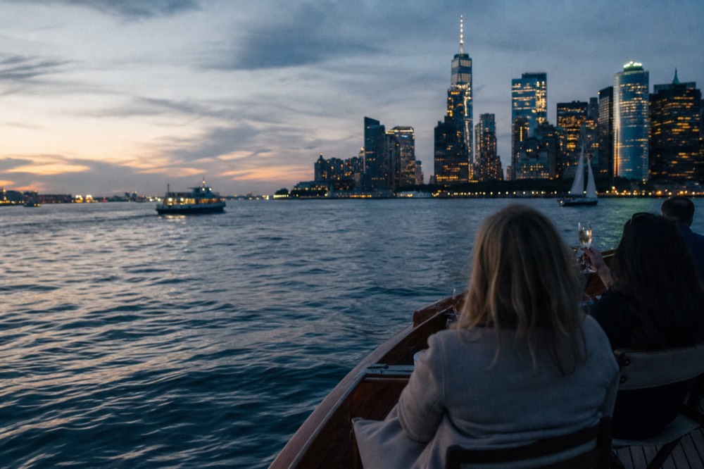 Passengers holding champagne flutes on the open teak deck of a small classic motor yacht at golden hour with the Manhattan skyline behind