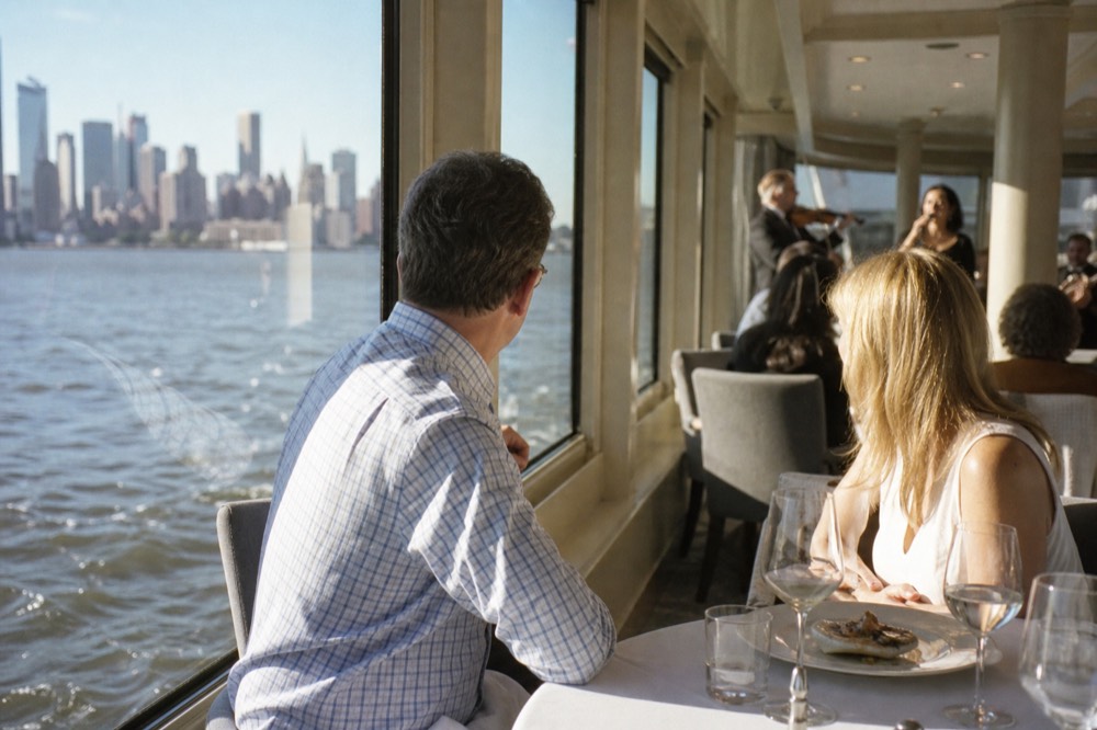 A couple seated at a window table in a glass-walled dining room on a cruise vessel, a violinist and singer performing in soft light behind them