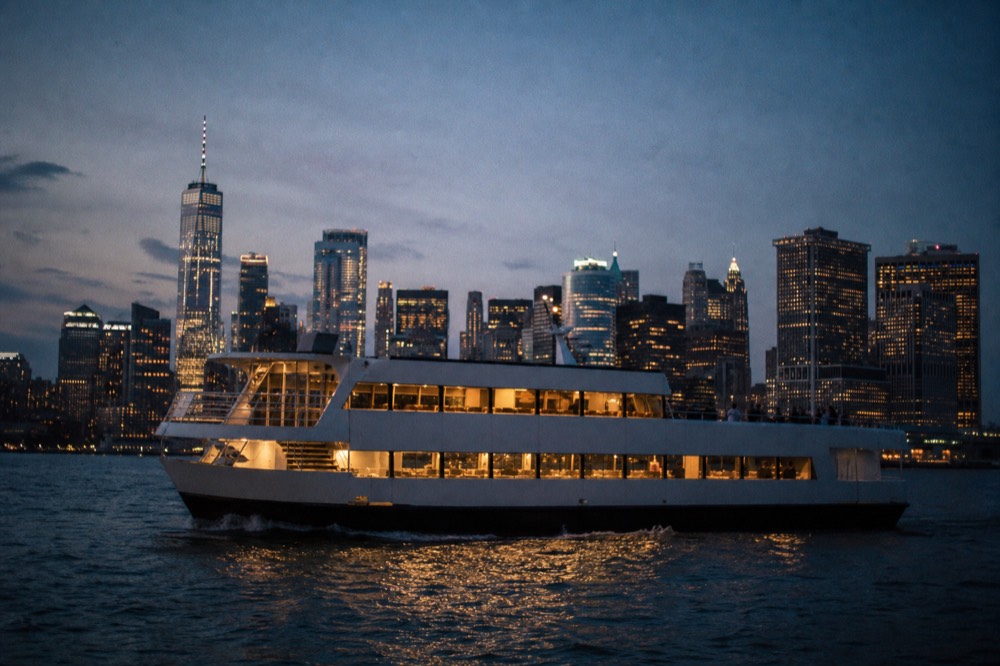 Wide view of a dinner cruise vessel on New York Harbor at blue hour, lit windows along both decks and the Lower Manhattan skyline behind it