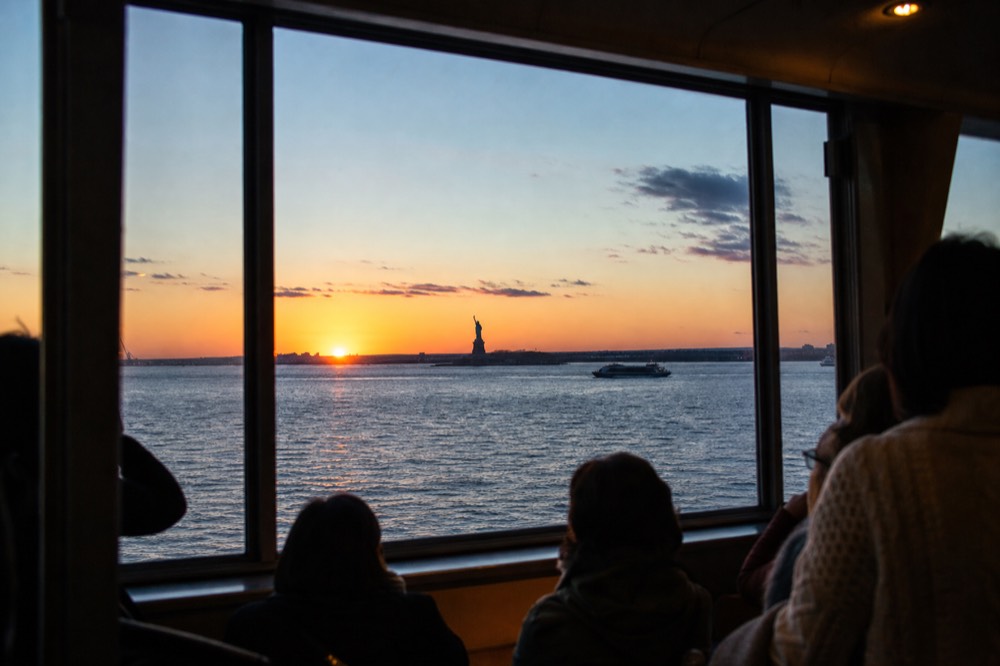 Wide view of New York Harbor at the moment of sunset with the sun touching the New Jersey shoreline and the Statue of Liberty in silhouette