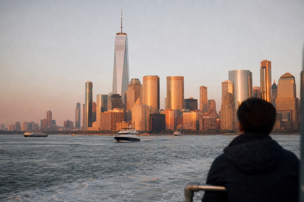 Low sun lighting the west faces of lower Manhattan towers in deep orange, viewed from harbor water with small boats in the middle distance