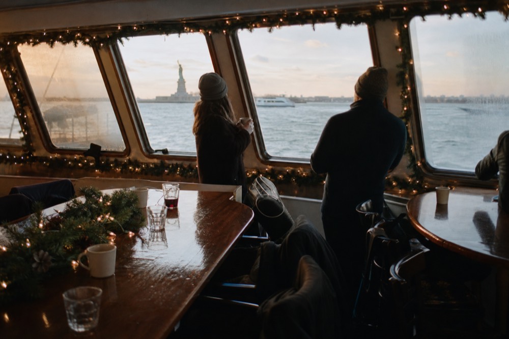 Interior of a small yacht observatory decorated with pine garlands and warm string lights, guests holding mugs beside frosted windows