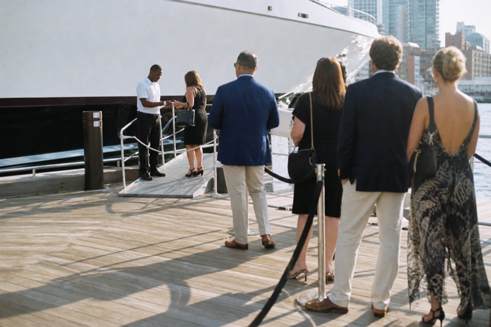 Crew member checking a boarding pass at the head of a gangway while a short queue of well-dressed guests waits behind a rope