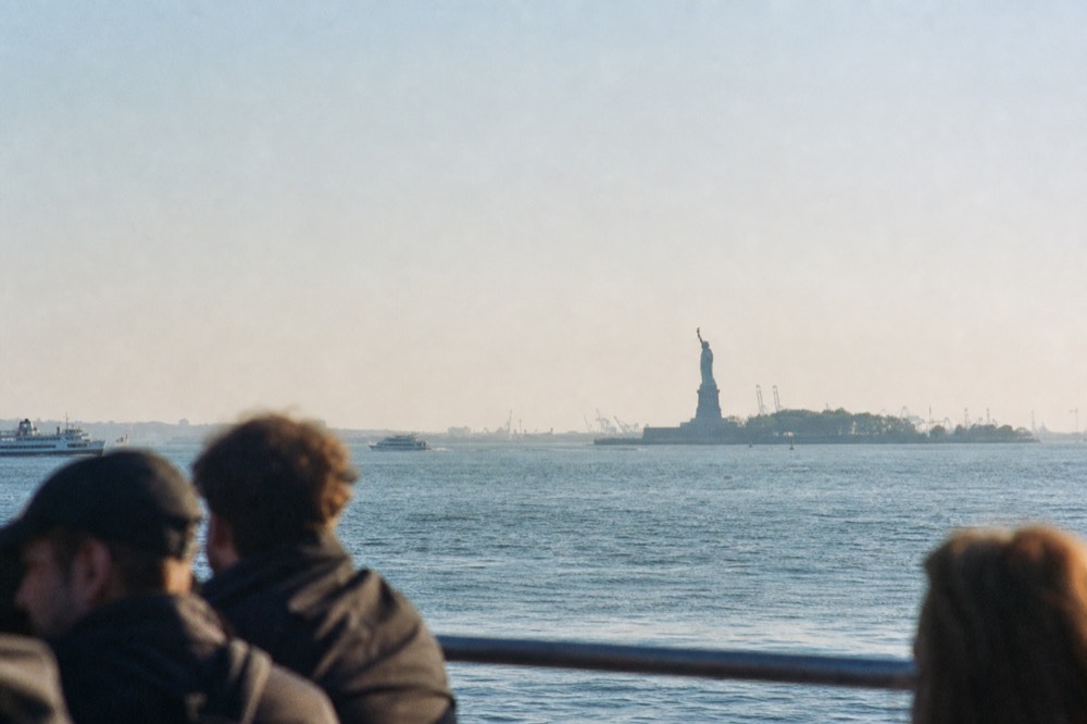 The Statue of Liberty seen at middle distance across open harbor water from a moving boat, small in frame against a pale sky