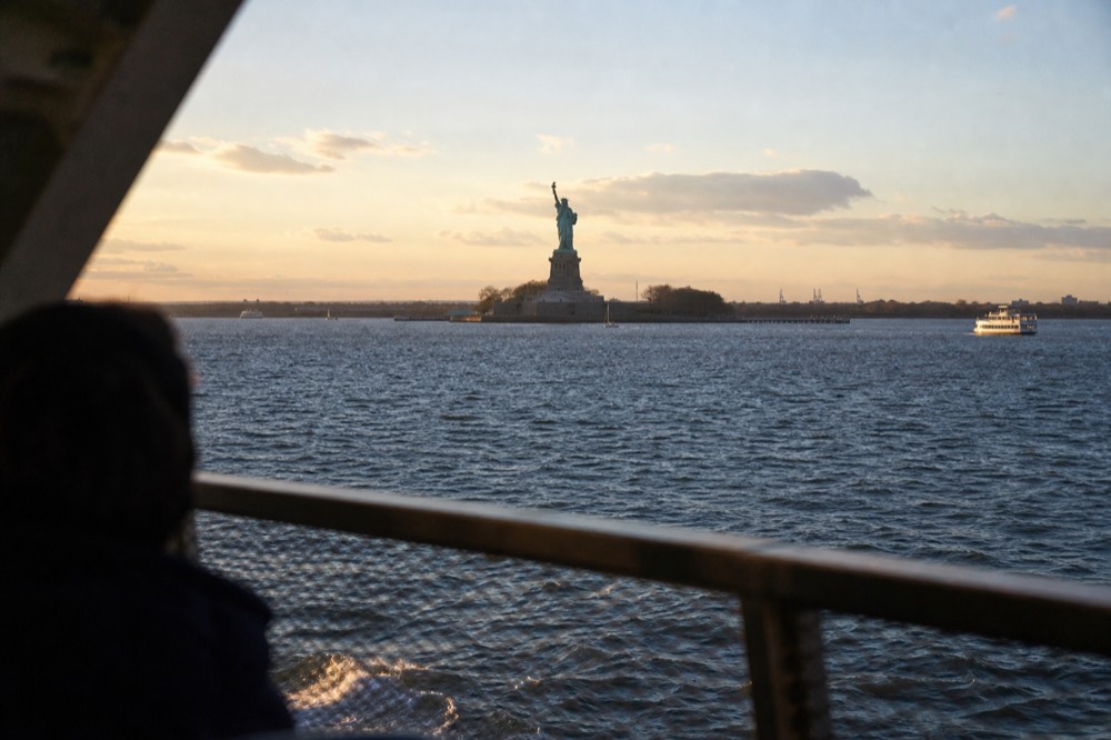 Statue of Liberty seen at a distance across open harbor water from the deck of a passing vessel, late afternoon light