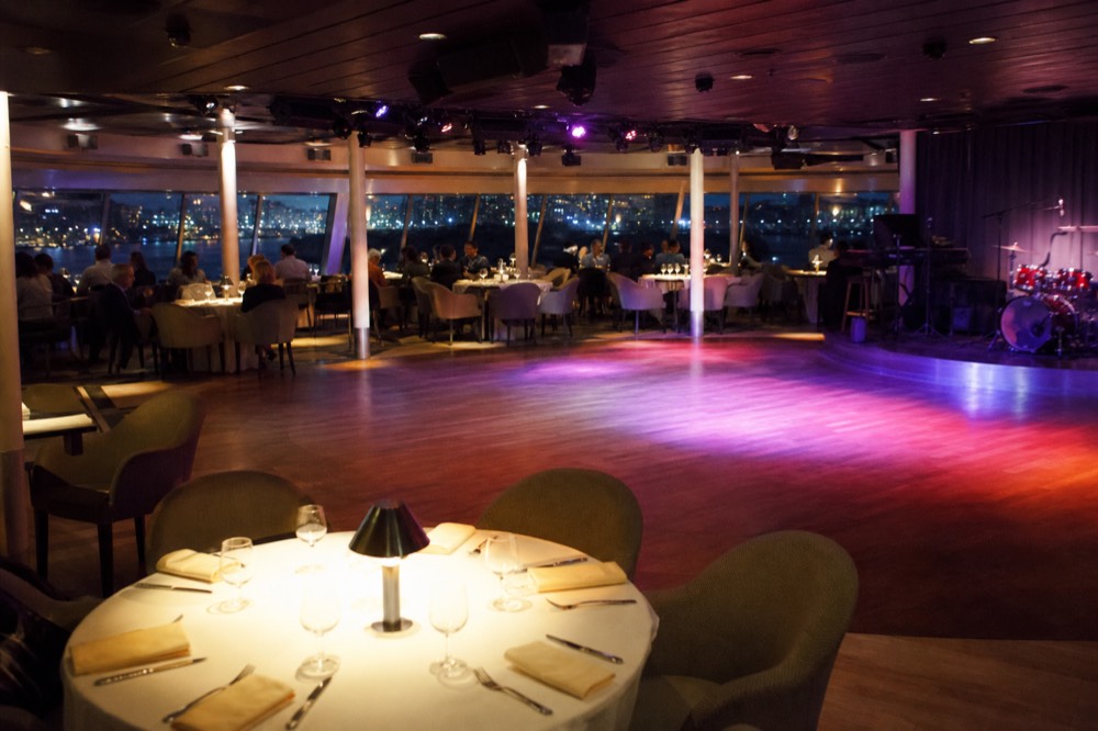 Interior deck of a large cruise vessel with round dining tables, a clear dance floor and coloured lighting, windows showing water at night