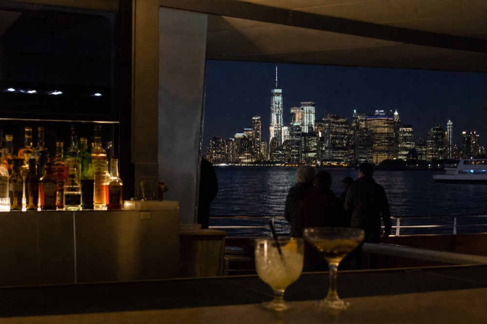 Onboard bar on a harbor cruise vessel with bottles backlit on shelves and two cocktail glasses on the counter, city lights beyond a window