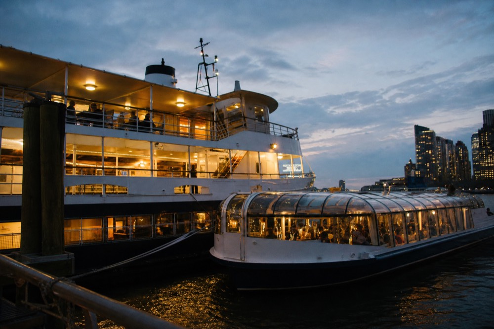 Two different harbor dinner vessels moored at adjacent berths in early evening, one tall with open decks, one low and glass-roofed