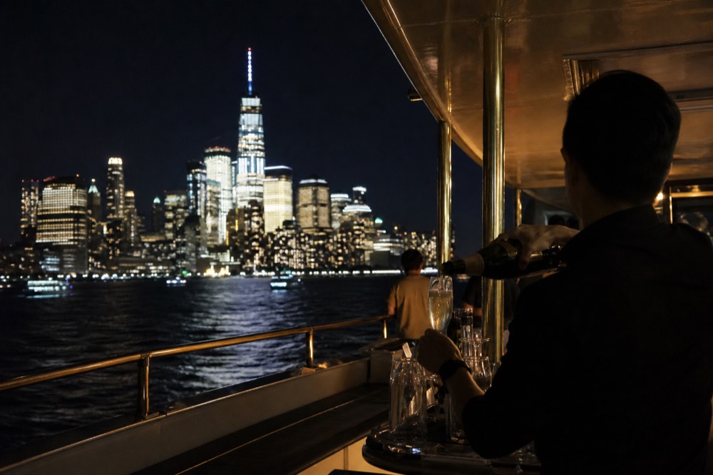 Bartender pouring sparkling wine into a flute at a small boat bar with brass rails and warm interior lighting